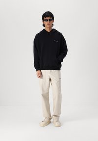 Hanorac negru cu buzunar frontal și un logo mic, asortat cu pantaloni bej și adidași albi, purtând ochelari de soare negri. Design minimalist.