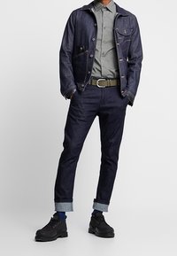 Jeansowa kurtka z kieszeniami, szara koszula, ciemnoniebieskie spodnie dżinsowe z podwiniętymi nogawkami oraz czarne buty na sznurowadła. Charakteryzuje się swobodnym, wytrzymałym stylem.