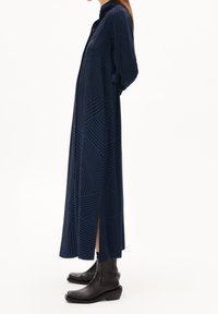 Kvinna i en lång marinblå klänning med geometriskt mönster och en sidoslit, iklädd svarta läderstövlar till mitten av vaden, stående i profil.