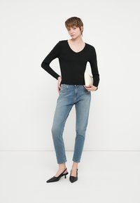 Zwarte lange mouwen V-hals top gecombineerd met lichtblauwe rechte jeans en zwarte puntige hakken. Het model houdt een crèmekleurige clutch vast.