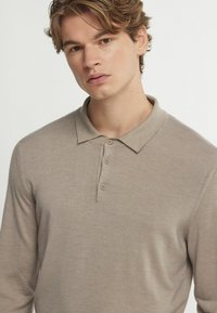 Beige långärmad polo skjorta i mjukt material med krage och tre knappknäppning. Har en slät yta och en figurnära design.