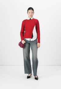 Vrouw in een rood asymmetrisch knopen trui, witte shirt met zwarte stropdas, grijze wijde jeans, zwarte hakken en een bordeauxrode tas staat tegen een witte muur.