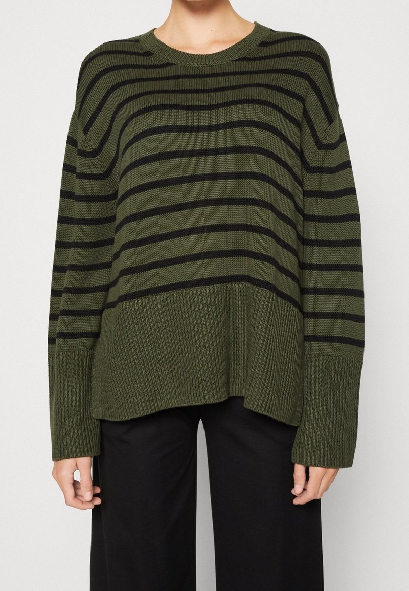 Personne portant un pull vert olive à rayures horizontales noires et larges poignets côtelés, associé à un pantalon noir ample.