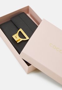 Portafoglio in pelle nera in una scatola rosa, con un lucchetto dorato e una finitura testurizzata. Il coperchio della scatola mostra "COCO" in lettere dorate.