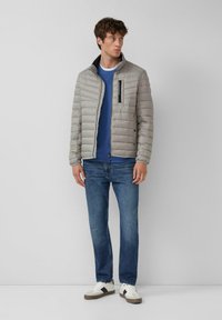 Lichtgrijze donsjas met een ritssluiting aan de voorkant en zwart pocketdetail, gecombineerd met een blauw shirt met lange mouwen, blauwe jeans en witte sneakers.