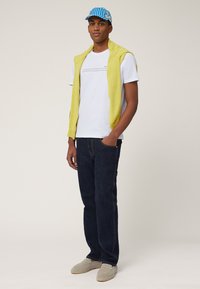 White t-shirt with graphic, yellow knit sweater draped over shoulders, dark denim jeans, and gray loafers. Blue striped cap completes the look.