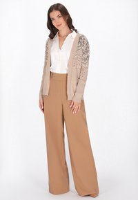 Beżowy kardigan z cekinowymi detalami na ramionach, połączony z kremową bluzką i beżowymi spodniami o szerokich nogawkach i wysokim stanie. Gładka faktura materiału.