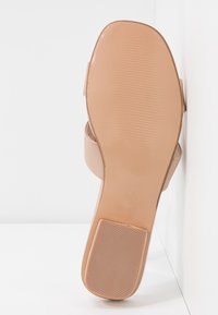 Semelle d'une sandale beige avec un talon carré texturé antidérapant et une semelle lisse, reposant contre un mur blanc et une surface blanche.