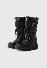 Botas de invierno impermeables negras con un diseño acolchado, cremallera en la parte frontal, cordones elásticos y suelas de goma con agarres texturizados. Acento de logotipo dorado.