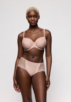 Vrouw met kort blond haar, gekleed in een lichtroze kanten beha en bijpassend hoog uitgesneden ondergoed met kanten rand, staand tegen een effen grijze achtergrond.