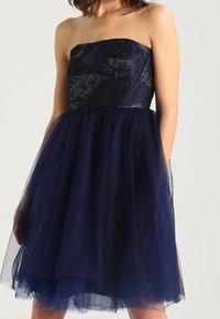 Axelös klänning i marinblått; har en spetsöverdel med blommönster och en lager på lager av tyllkjol. Mjuk textur och knälång design.