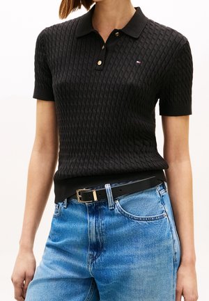 Femme portant un polo noir texturé à manches courtes, rentré dans un jean taille haute bleu clair avec une ceinture noire.