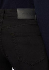 Černé džínové kalhoty s texturovanou koženou etiketou s nápisem "TRUSSARDI." Obsahují standardní kapsy a nenápadné detaily prošívání.