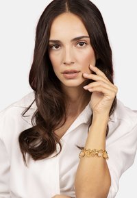 Pulsera dorada con cuentas redondas y translúcidas y eslabones, llevada en la muñeca de una persona. El fondo es blanco liso.