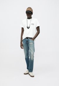 Weißes T-Shirt mit kurzen Ärmeln und grafischem Text, hellblaue Jeans, beige Sneaker und eine mehrfarbige gestrickte Mütze. Schwarzes Accessoire um den Hals.