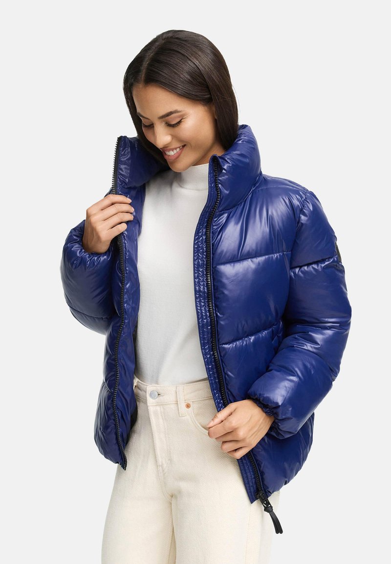 Granatowa kurtka puchowa, błyszczące wykończenie, wysoki kołnierz, zamek z przodu, elastyczne mankiety, noszona na białym swetrze, w połączeniu z kremowymi spodniami.