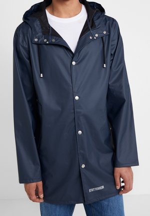 Homme portant un imperméable à capuche bleu foncé avec des boutons-pression par-dessus une chemise blanche et un jean bleu.