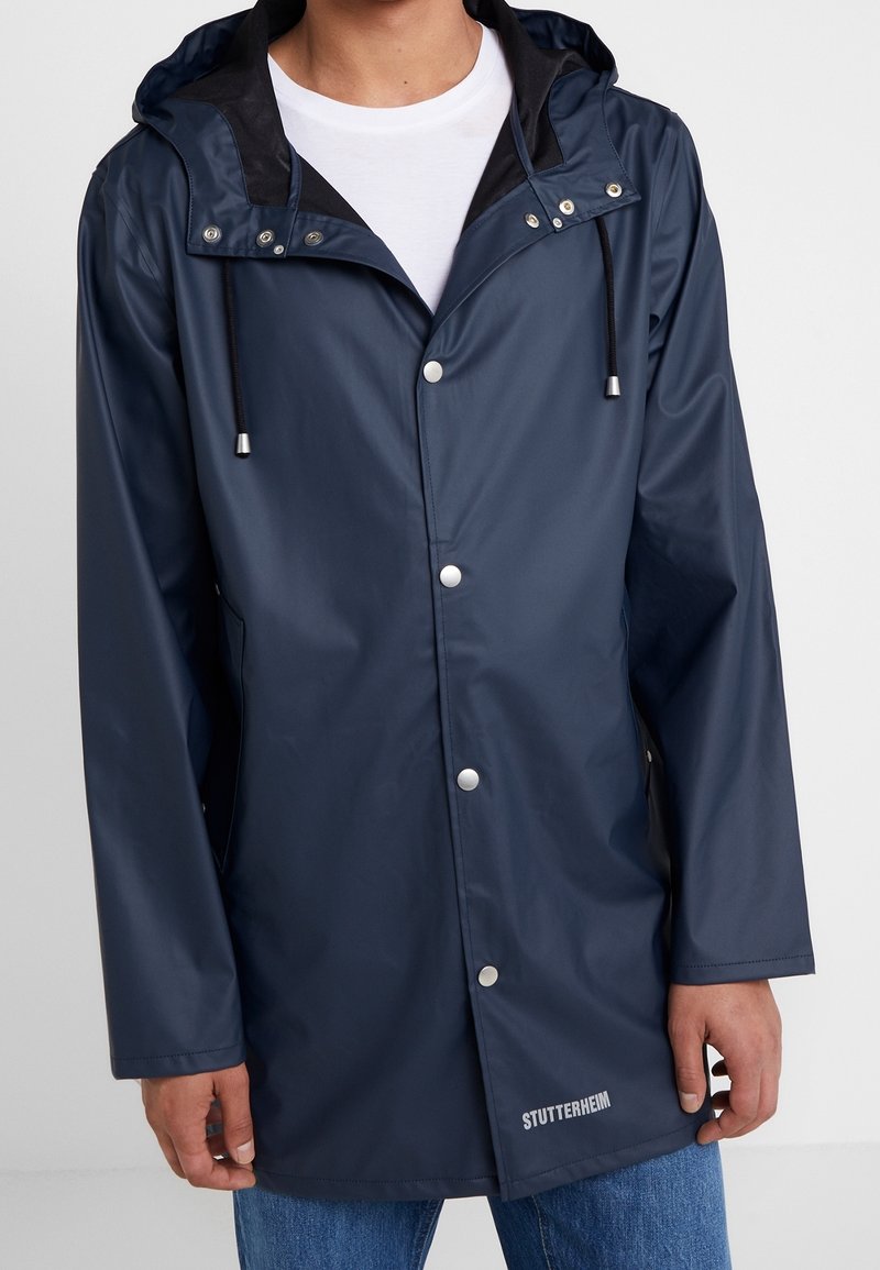 Homme portant un imperméable à capuche bleu foncé avec des boutons-pression par-dessus une chemise blanche et un jean bleu.