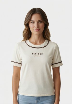 Donna con capelli castano ondulati che indossa una T-shirt a maniche corte color crema con bordi marroni e scritta "YES ZEE", abbinata a jeans azzurri chiari.