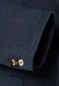 Donkerblauwe, gestructureerde stof van een mouw met vier knopen: twee zwarte, één donkerbruine en één crèmekleurige. Zichtbare stiksels langs de rand van de manchet.