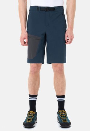 Homme portant un short bleu marine avec une poche latérale noire, une ceinture noire, des chaussettes noires à rayures blanches, et des chaussures de randonnée bleues avec des lacets jaunes.