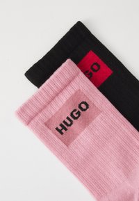Pair of ribbed socks, one pink with black "HUGO" logo on cuff, one black with red and black "HUGO" logo, laid overlapping on white background.