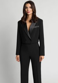 Czarna krótka marynarka z satynowymi szpiczastymi klapami, podwójnymi guzikami i dopasowanym krojem, zestawiona z dopasowanymi spodniami. Gładka faktura, formalny styl.