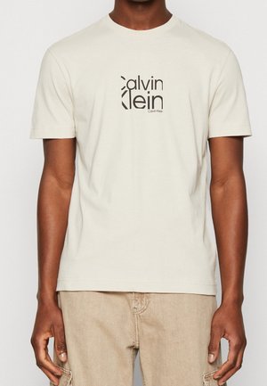 Homme portant un t-shirt Calvin Klein blanc cassé avec un logo noir, associé à un pantalon beige clair.