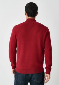 Red knitted sweater with a high collar, ribbed cuffs, and hem, viewed from the back. Fabric appears smooth with a slight sheen.