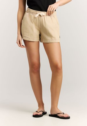 Persoon draagt beige gestructureerde shorts met elastische tailleband en strik, zwarte top en zwarte teenslippers, staand tegen een neutrale achtergrond.