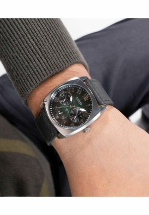 Quadratische Uhr mit schwarzem Zifferblatt, Gehäuse in Silberfarbe und großen Ziffern. Schwarzes Lederarmband mit strukturierter Oberfläche. Silberne Krone an der Seite.