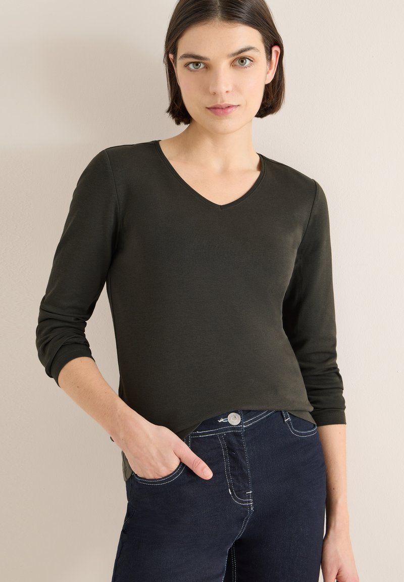 Blusa verde scuro a maniche lunghe con scollo a V, realizzata in tessuto morbido. Presenta maniche aderenti e un orlo ampio, abbinata a jeans scuri.