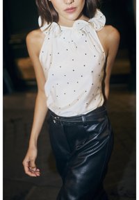 White sleeveless top with black polka dots and a ruffled collar, paired with black perforated leather high-waisted pants and a belt.
