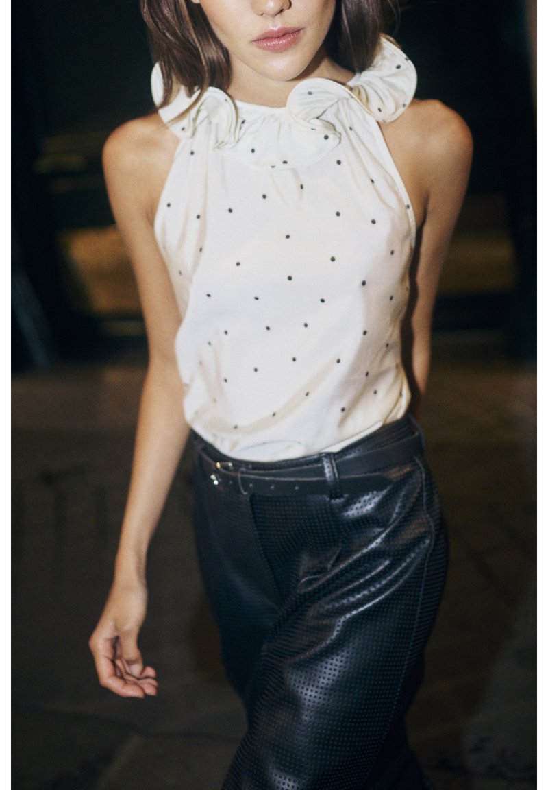 White sleeveless top with black polka dots and a ruffled collar, paired with black perforated leather high-waisted pants and a belt.