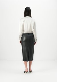 Femme de dos portant une blouse blanche à manches longues, une jupe mi-longue en cuir noir avec une fente à l'arrière, et des mules à talons noirs sur fond blanc.