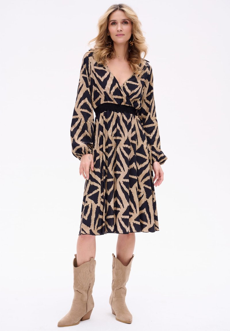 Maxi-jurk met een V-hals, lange mouwen en een zwart-beige abstract patroon. Heeft een gerimpelde taille en is gecombineerd met tan cowboylaarzen.