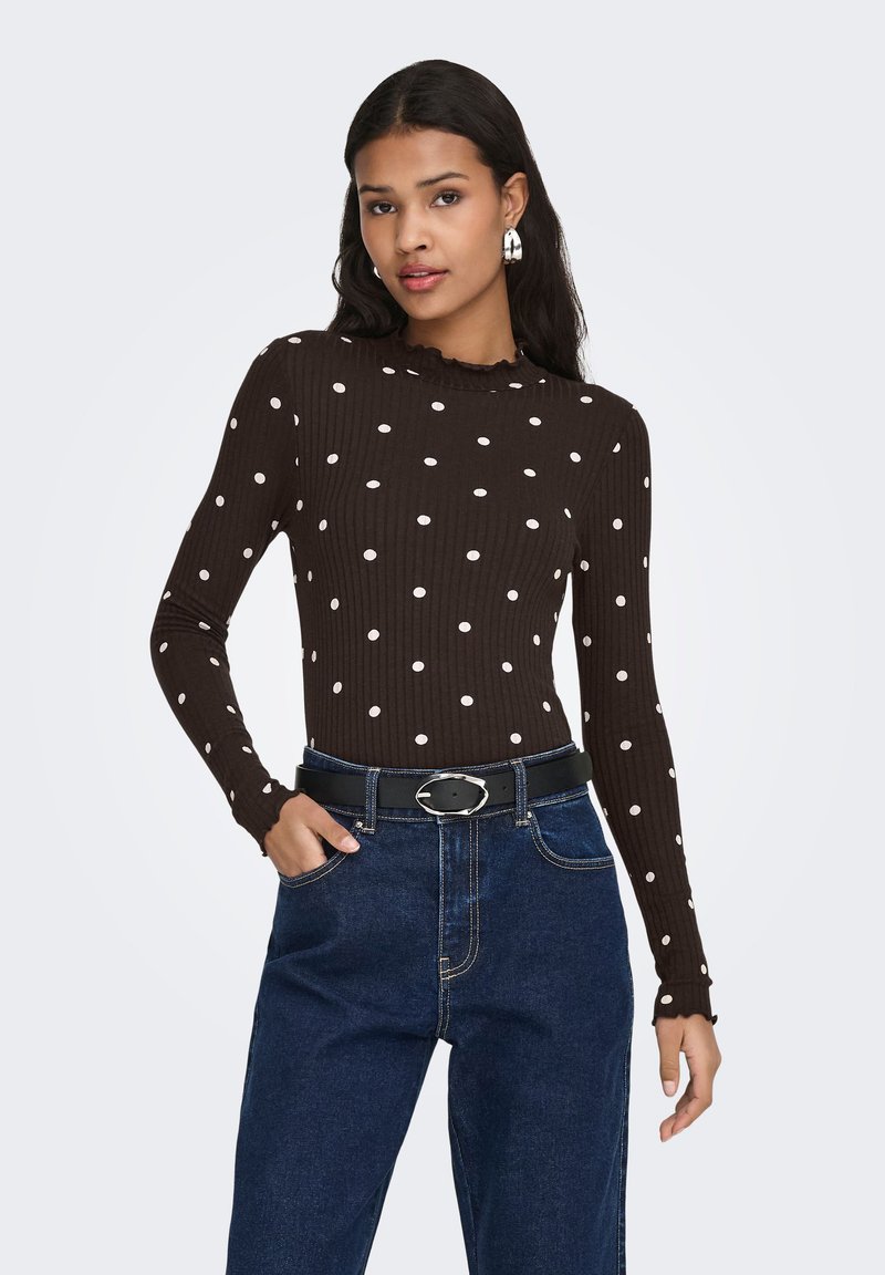 Brun ribstrikket langærmet top med hvide polkaprikker og en flæsekant ved halsen, kombineret med mørkeblå højttaljede jeans og et sort bælte.