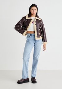 Bruine neplederen jas met crèmekleurige fleece voering, knoopsluiting en stikdetails, gecombineerd met lichtblauwe jeans en zwarte platform schoenen.
