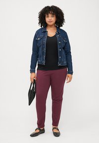 Ženska s kodrastimi lasmi nosi jeans jakno, črn top in maroon hlače, drži črno torbo. Stoji pred enobarvno ozadjem.