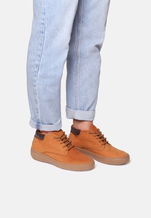 Brown leather high-top shoes with brown laces and a rubber sole, paired with rolled-up light blue jeans, revealing shoe design details.