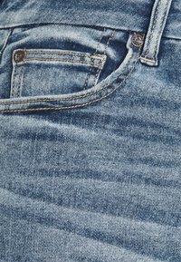 Siniset farkut, joissa on vaalea pesu, näkyvät saumat ja pieni etutasku metallisella napilla. Kankaan tekstuuri esittää haalistunutta kuviota.