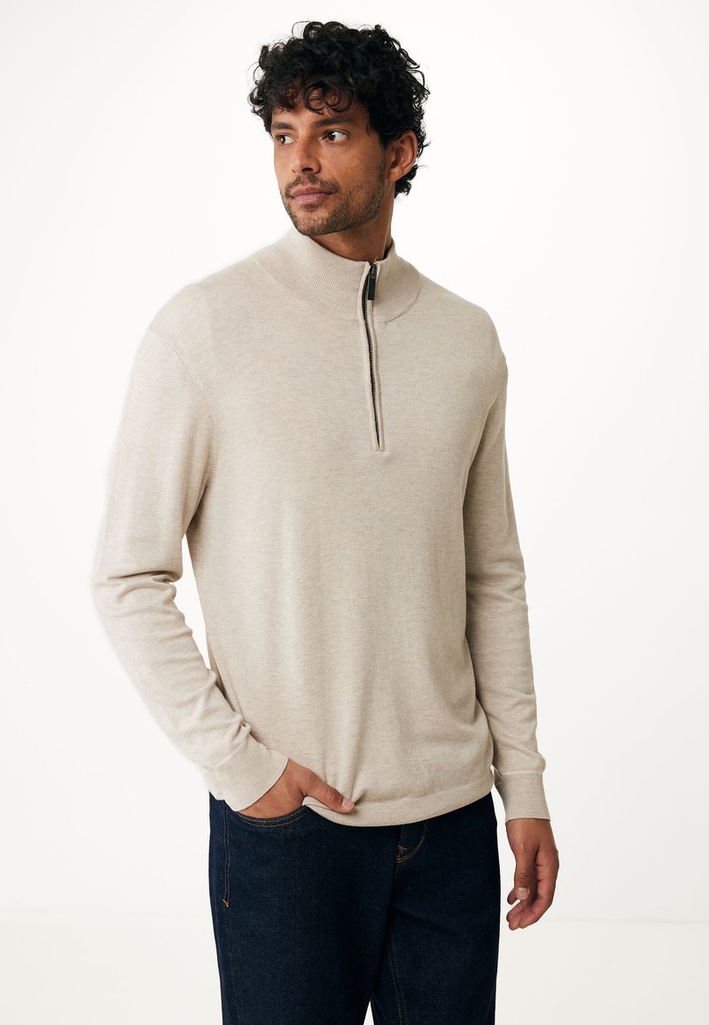 Beżowy sweter z półzamkiem, z długimi rękawami, wykonany z miękkiego materiału. Posiada wysoki kołnierz oraz minimalistyczny design. Noszony z ciemnoniebieskimi dżinsami.