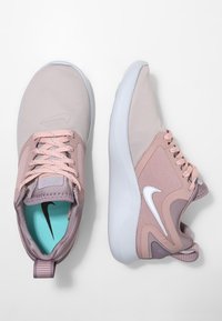 Scarpe Nike in rosa chiaro e grigio, realizzate in rete e materiali sintetici. Le caratteristiche includono un design con lacci, un collare imbottito e accenti bianchi.