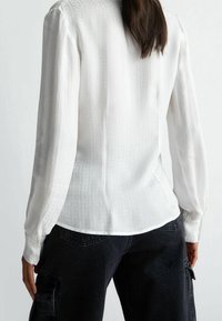 Blusa bianca a maniche lunghe con un motivo testurizzato sottile, polsini abbottonati e orlo curvo, abbinata a pantaloni cargo scuri.