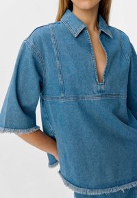 Chemise en denim bleu clair, avec une demi-buttonnière, des manches courtes et un ourlet effiloché. Une couture distinctive traverse le devant.