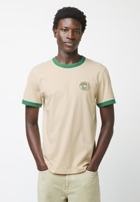 Beige t-shirt met groene geribbelde ronde halslijn en mouwen. Beschikt over een cirkelvormig logo op de borst. Zacht katoen materiaal, casual pasvorm.