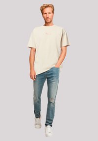 Beige katoenen T-shirt met korte mouwen en rode tekst, gecombineerd met lichtblauwe versleten jeans. Schoeisel: witte sneakers.