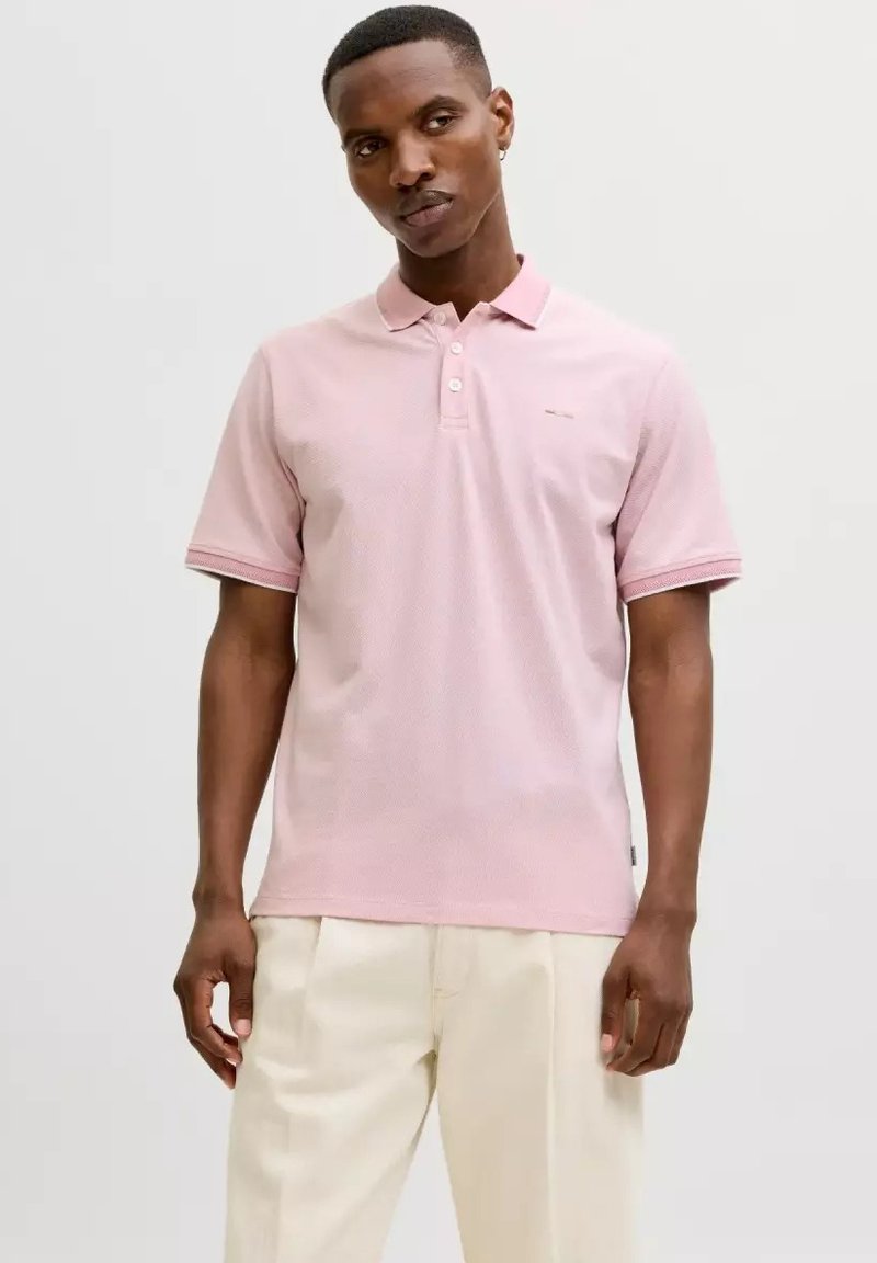 Mann trägt ein hellrosa kurzärmeliges Poloshirt und lockere cremefarbene Hosen vor einem einfarbigen hellen Hintergrund.