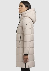 Kerge beeži tooni puffer-jakk, millel on kapuuts, horisontaalne õmblus, mustad tõmblukud, reguleeritavad nöörid ja säärevarde reguleeritavad varrukad.