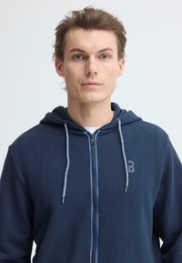 Sweat à capuche zippé bleu marine avec tissu texturé, cordons gris, et petit logo sur la poitrine gauche. Comprend une capuche et une fermeture éclair frontale.